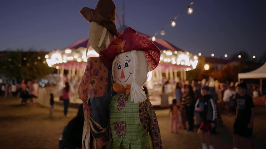 Colorful scarecrows decorate a fairground as people walk around in the background in the amusement park at dusk