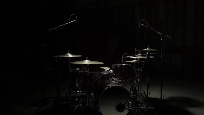 In a dark room a weak light shines upon a drum set with several cymbals which casts shadows on the featureless ground