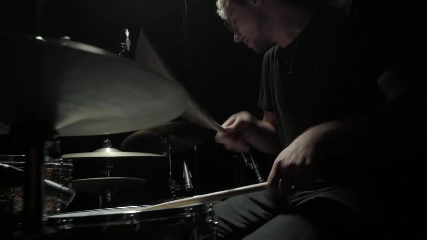 A young man is sitting at a drum set in a darkened room and playing the drums with great speed and vigor