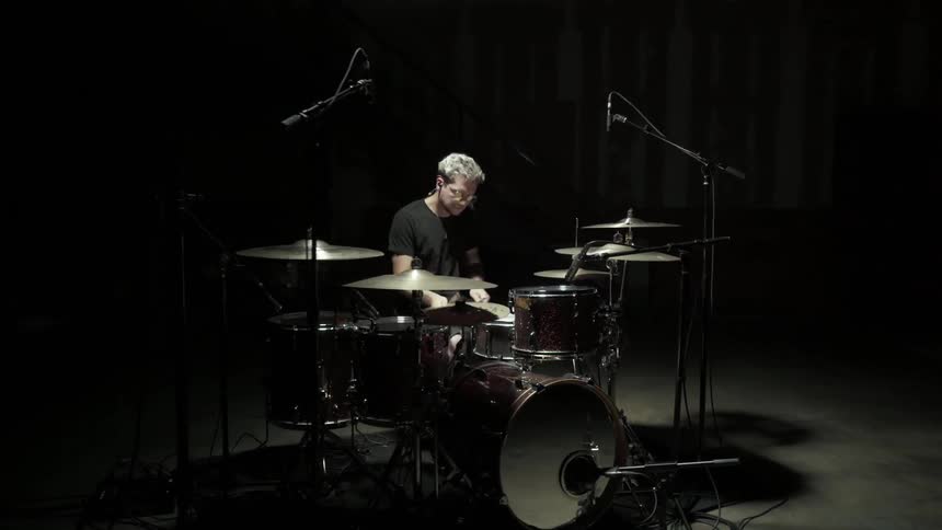 A man wearing glasses sitting on a dark stage by himself is drumming intently on a set of drums and pauses for a brief second to push the glasses up on to the bridge of his nose