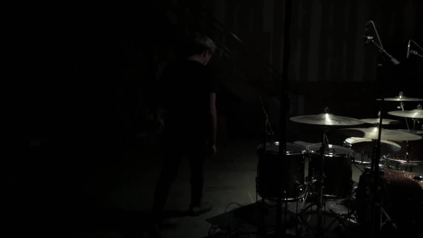 Man goes to the drum set in the spotlight and sits down in a dark room