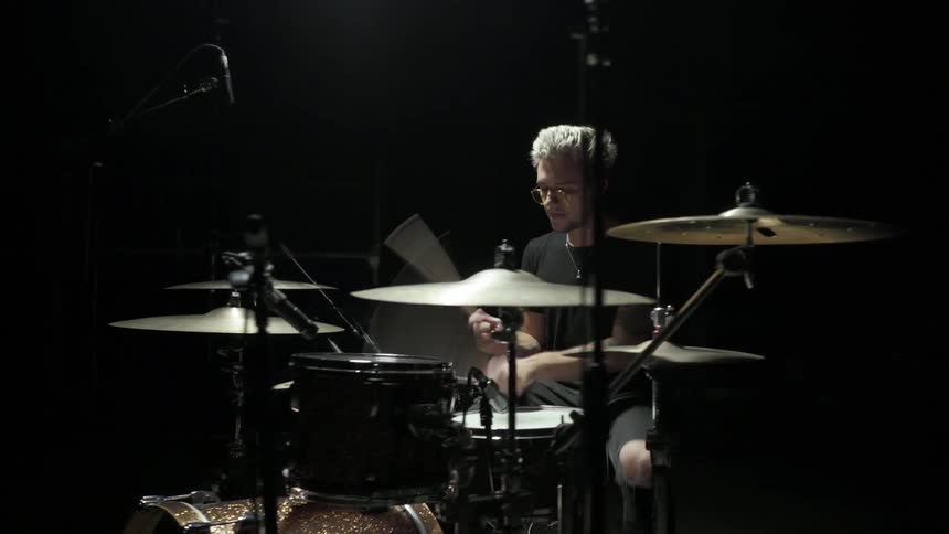 A blonde man wearing glasses vigorously plays a drum kit