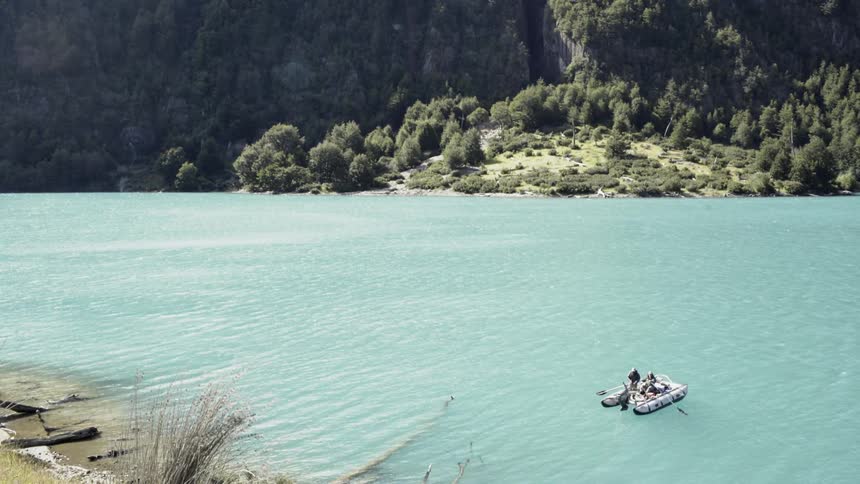 Two fishermen fish on a boat floating on turquoise lake with the view of steep mountain in the daytime