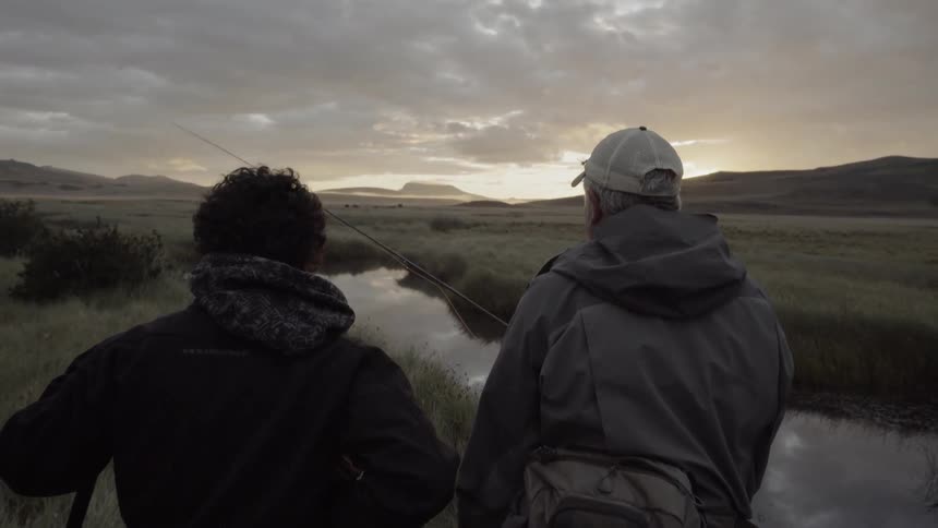 Two men holding fishing line stand near a river look at the horizon at sunset