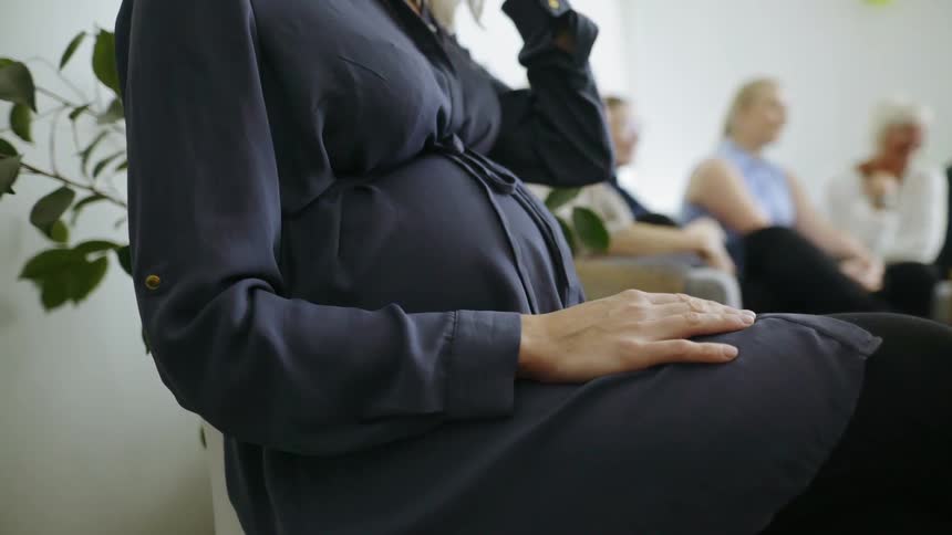 A pregnant woman sitting in a chair in a room with other women raises her hand to fix her hair