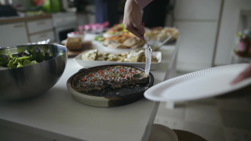 A woman holding a paper plate is cutting a piece of quiche using the edge of a pastry server and then scoops a piece up to put it on her plate