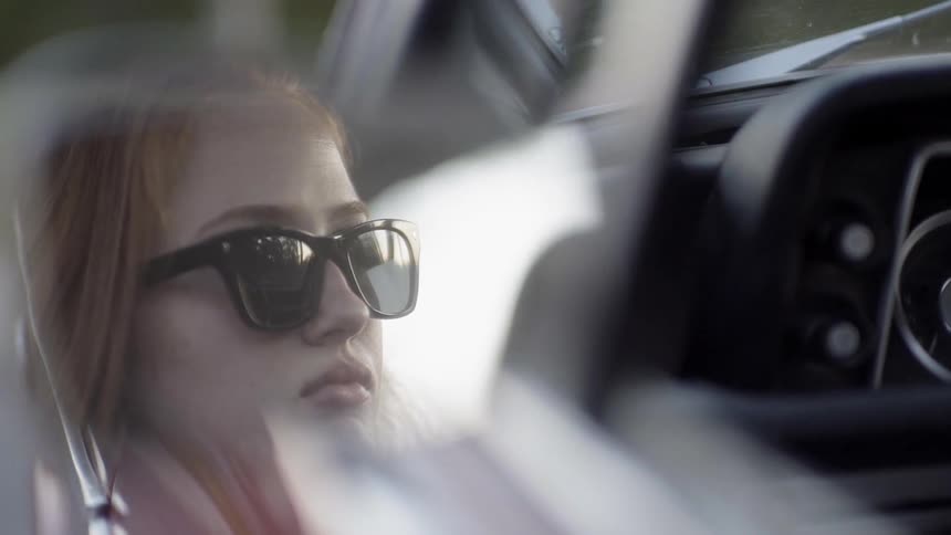 Ginger hair woman wearing sunglasses sits in the car and looks ahead in the daytime