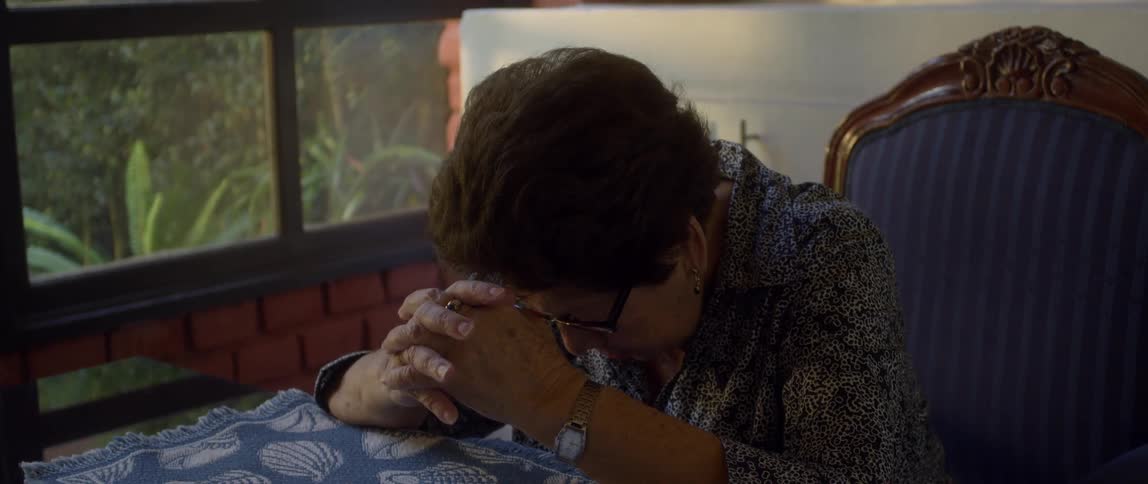 An older woman is sitting at a table inside with her head down and hands locked
