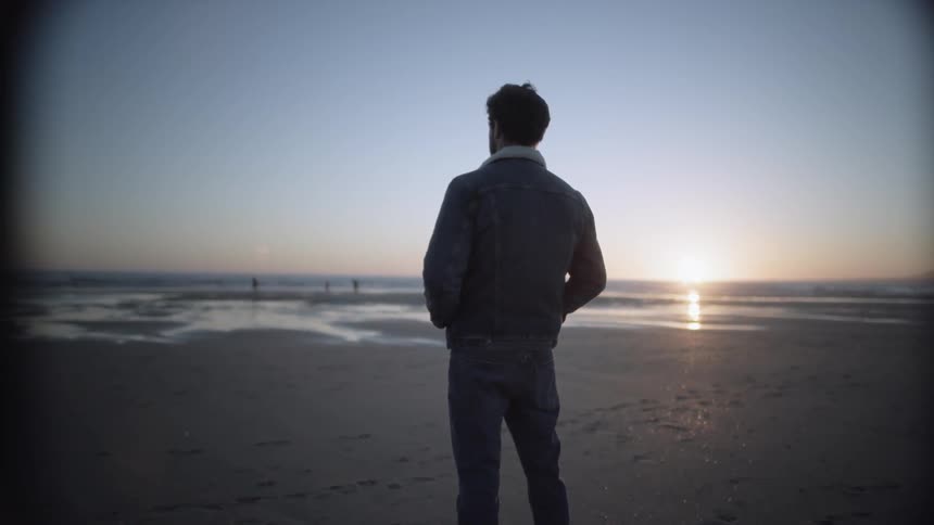 Man stands on the wet sand beach and looks at the sea while the sun goes down