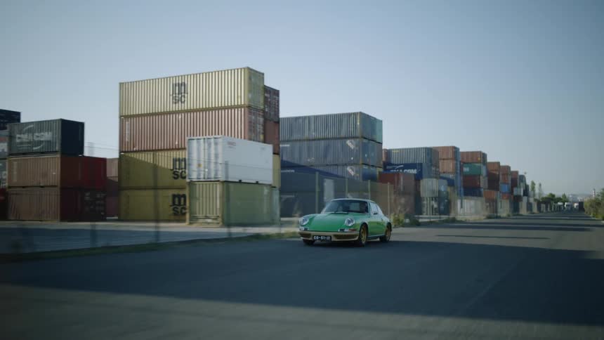 A green sports car travels on a road passing stacked shipping containers on a sunny day