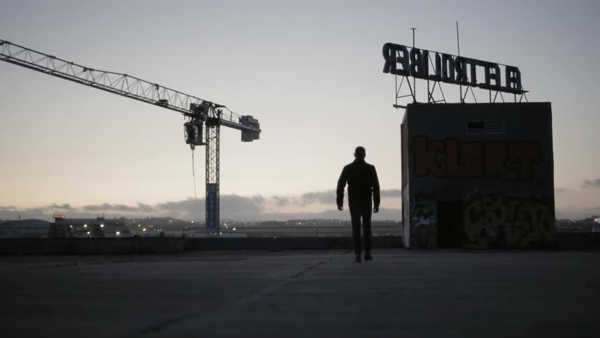A man walks along the roof of a tall building with confidence as large crane can be seen nearby amidst this urban area