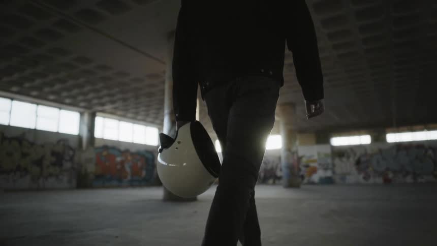 Man wearing denim jacket carries motorcycle helmet and walks indoors during daylight