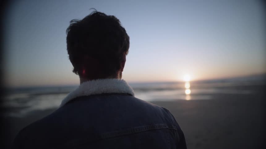 A man in a denim jacket stands on a beach at sunset