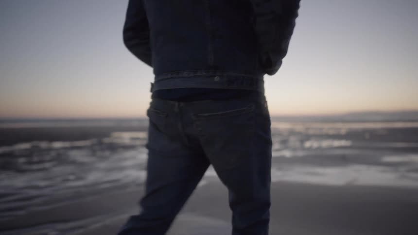 Man wearing denim walks in the wet sand at sunset