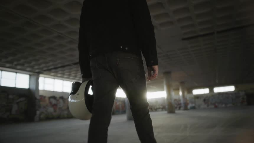 A man walks through an empty structure with columns and graffiti carrying his motorcycle helmet