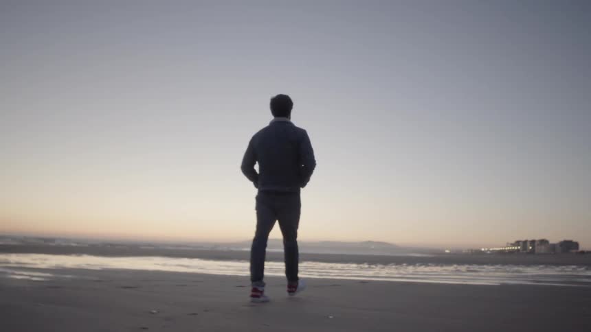 A man walks on the wet sandy beach at sunset with his hands in his pockets