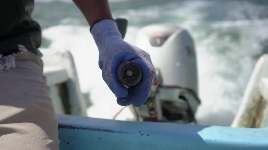 An man sits in a motor boat and while wearing a blue glove on his hand he holds the steering rod of the boats vibrating motor and subtly changed the direction of the boat