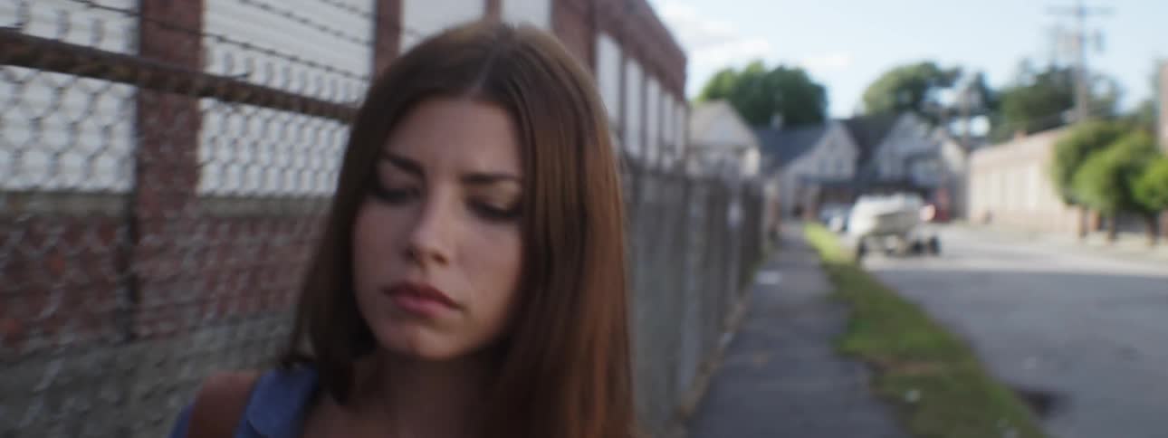 A young, brunette woman walking next to a fence when the view cuts away to the street