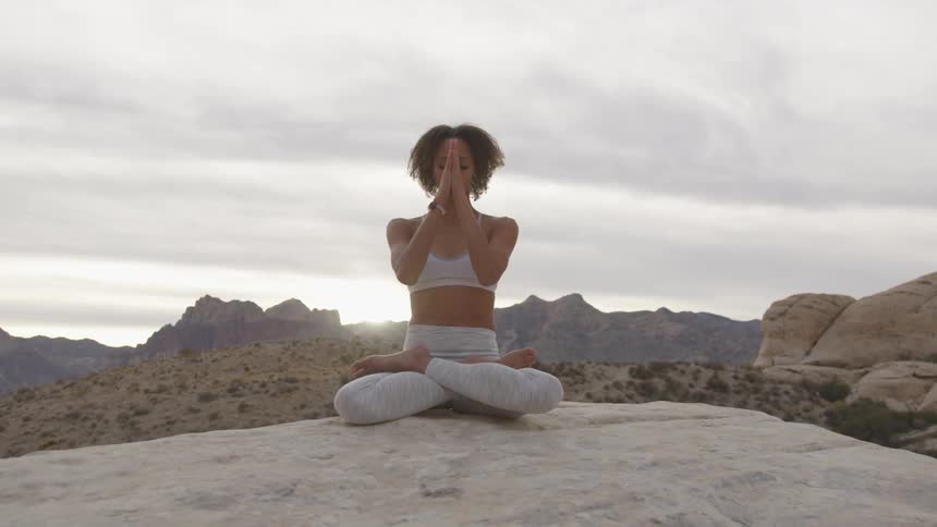 A woman sits on a rock in a desert with her hands pressed together and pulling them down in a yoga fashion