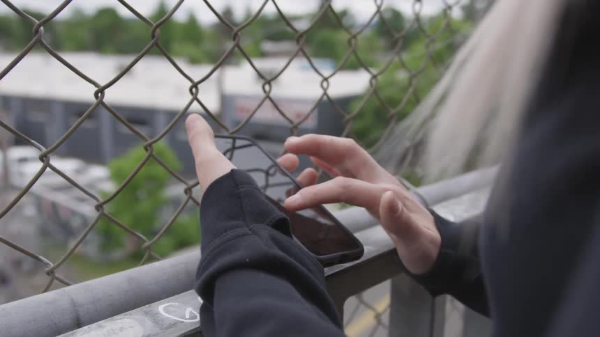 Blonde woman puts her hand on banister and uses smartphone in front of the wire fence on a cloudy day