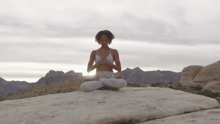 Brunette woman sitting cross legged and folding hands on a big rock performs yoga on a cloudy day