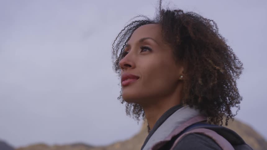 A woman is standing in front of a rocky hill and smiling towards the sky