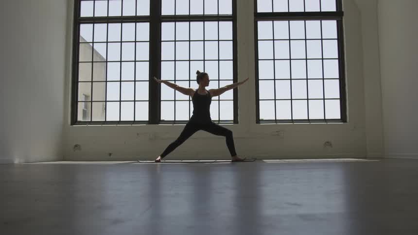 A woman does a yoga pose in front of big windows