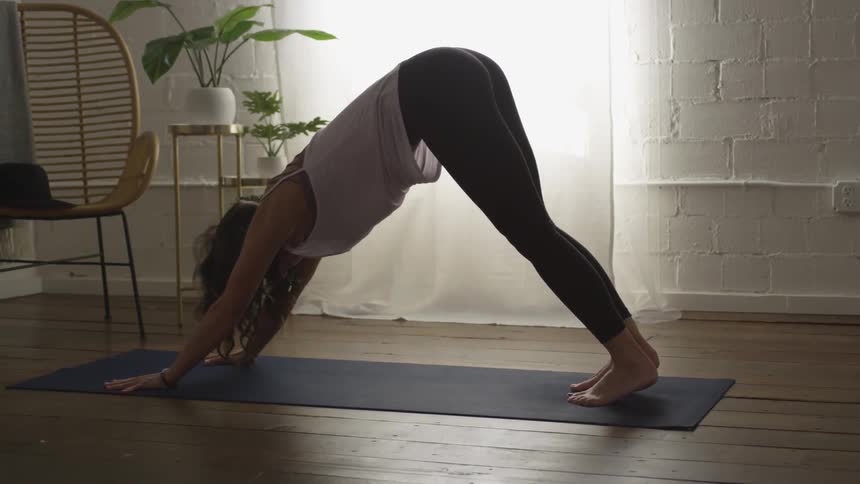 A thin woman in yoga pants and a loose tank top has her hands and feet both touching the ground as she performs the downward dog yoga pose