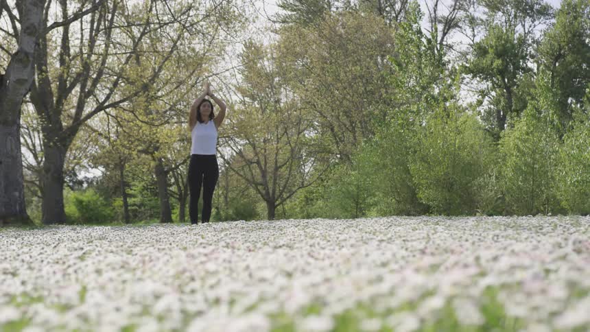A woman stretches her hands over the head with her palms together while standing in a field of flowers