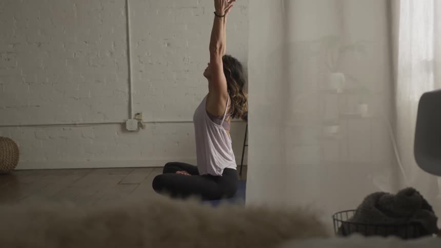 A woman is sitting on a yoga mat with her legs crossed as she stretched her arms up
