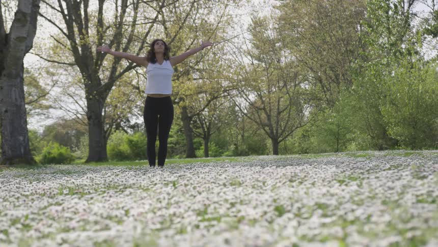 A woman in black yoga pants and a white tank top first reaches down to her toes and then up towards the sky as she stretches in a flower filled field amidst a forested area