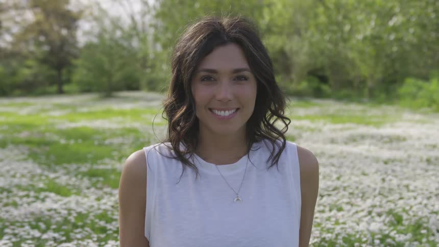 A woman is smiling as she stands in a field of grass and flowers