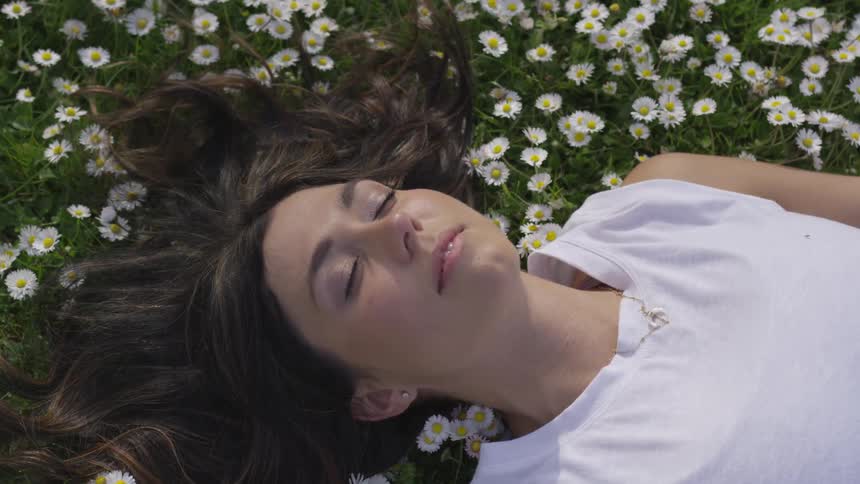 A brunette white woman lies in a field of daisies with her eyes closed