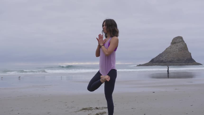 A woman is standing on the beach in a one leg yoga pose with her palms together