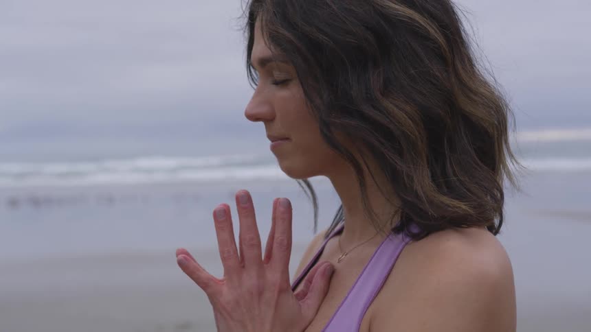 A woman is standing on a beach with her eyes closed and palms together with her fingers spread out