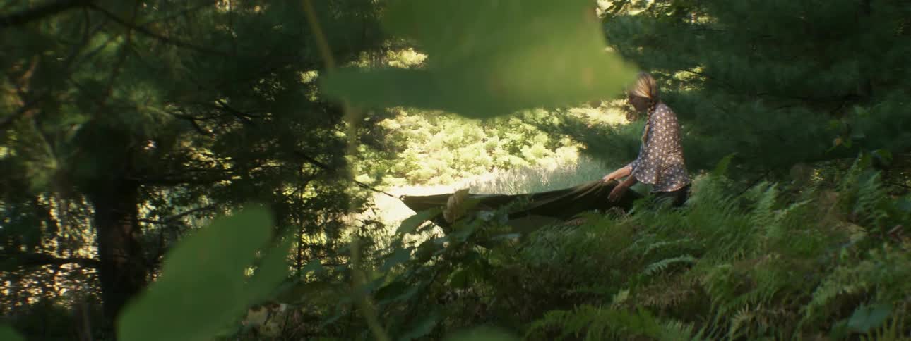 A blonde woman unraveling a hammock in a wooded area