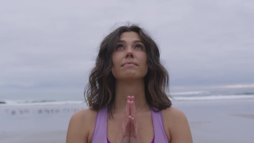 Woman folds her hands and looks up on the beach next to the sea on a cloudy day