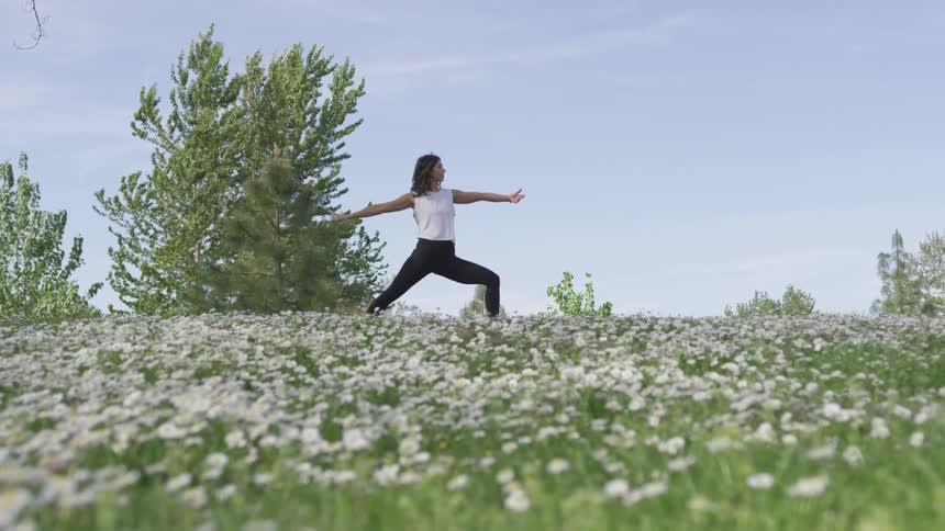 A woman in yoga pants and a sleeveless top performs a yoga pose while standing at the top of a hill covered in wildflowers