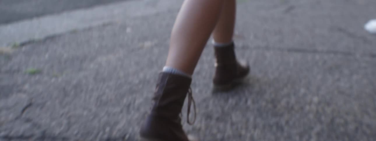 View of a woman's brown boots as she walks down a cracked, weed-filled sidewalk in an urban area