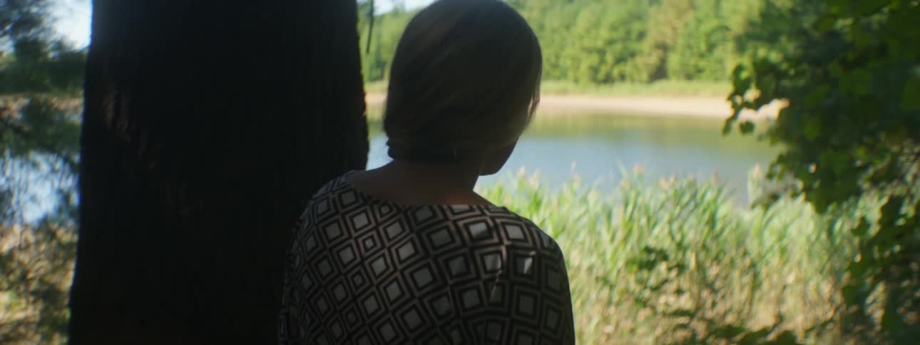 A blonde woman walking through some trees and brush towards a body of water