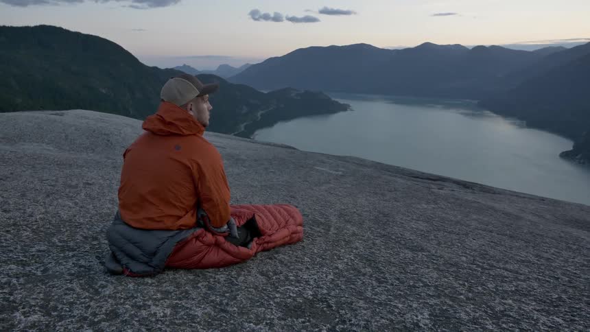 A man sitting in a sleeping bag on a cliff overlooking a body of water in the mountains