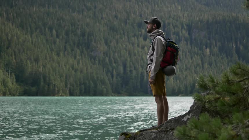 A young, bearded man wearing a red backpack stands on a shoreline looking at his surroundings