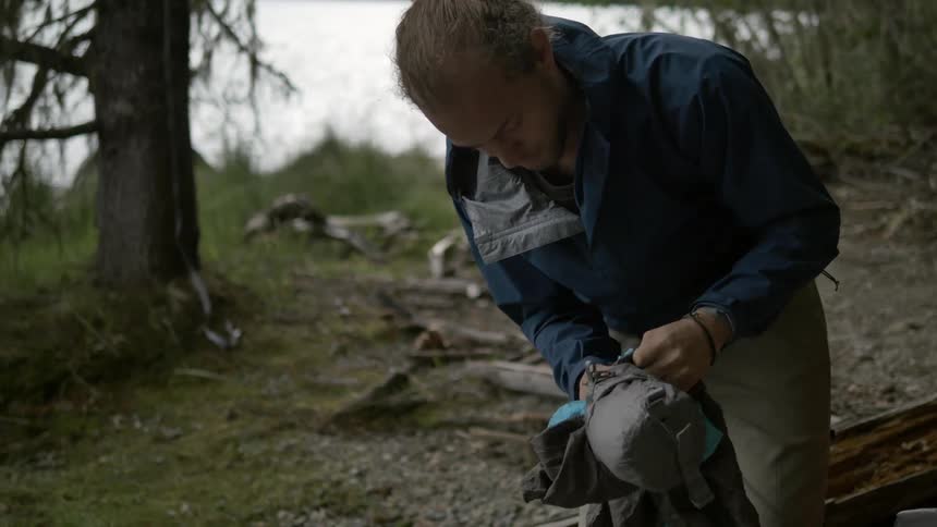 Man stuffs something into a small sack outside in the woods next to a lake