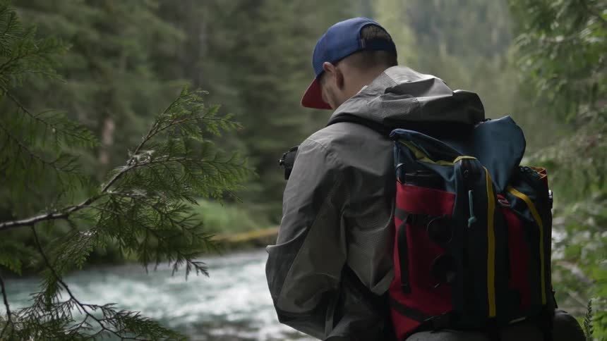 A ma checking the screen on his camera and making adjustments as he stands next to a mountain river
