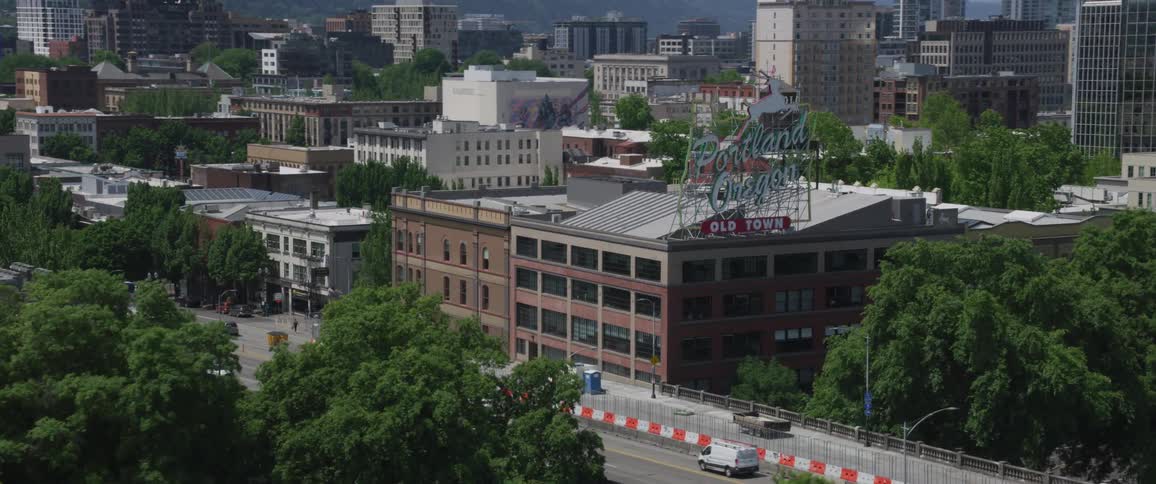 On a bright sunny day a large city with many trees can be seen while a prominent sign that displays portland oregon sits atop the edge of a building