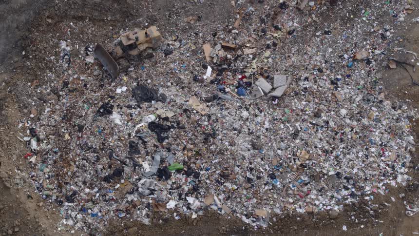 A bulldozer drives through a large trash landfill