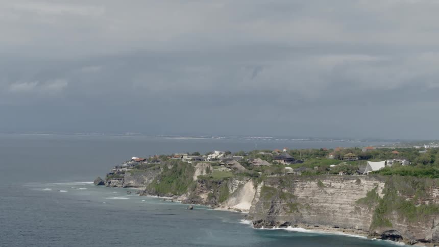 The camera flies along the coast with houses on top of a cliff