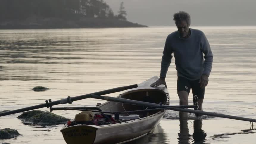 An old man walks slowly out of a lake while holding his right hand on the edge of a small boat with two oars