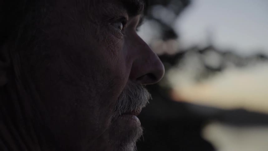 A mustachioed man with weathered and wrinkled skin turns his head and looks out over the landscape as the sun is below the horizon
