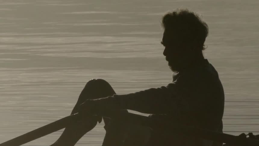 A man sits on a boat in the water with oars in his hands